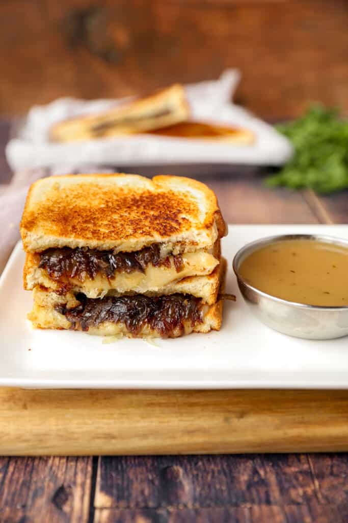 two stacked slices of French onion grilled cheese on a white plate with a silver dish of dipping broth.