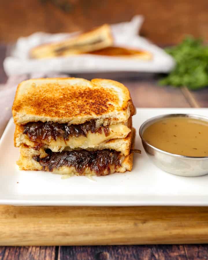 two stacked slices of French onion grilled cheese on a white plate with a silver dish of dipping broth.