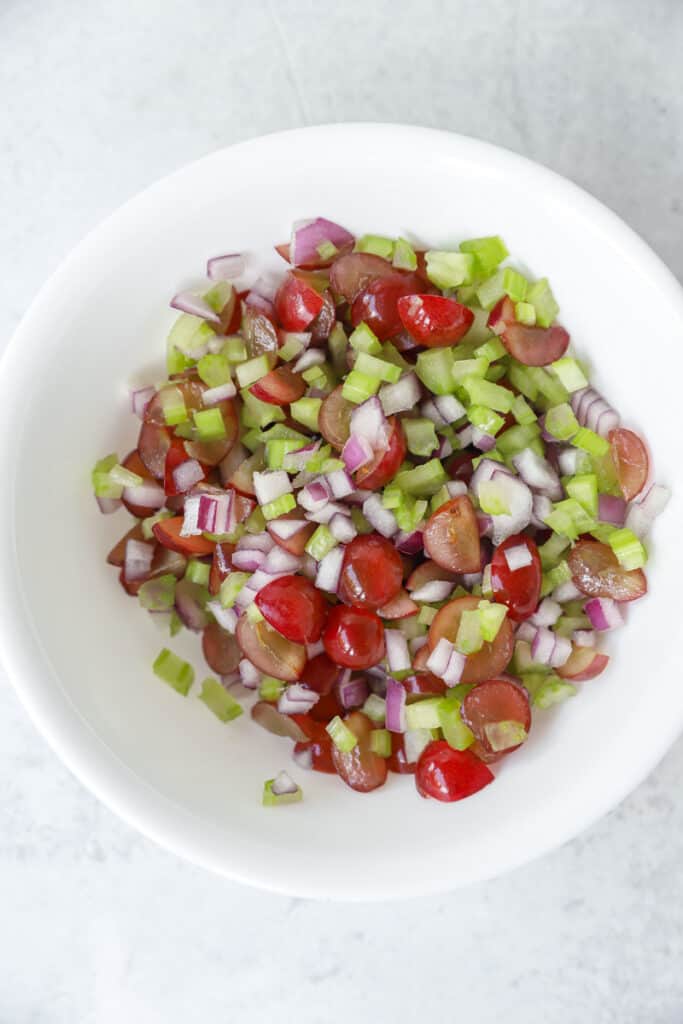 overhead photo of diced celery and red onions, and sliced red grapes in a round white bowl.