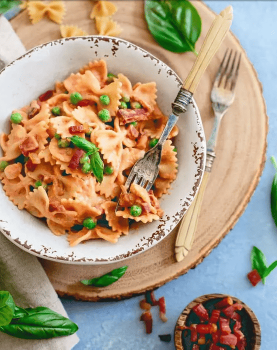 a bowl of the creamy tomato pasta with peas and a fork on a circular wood board.