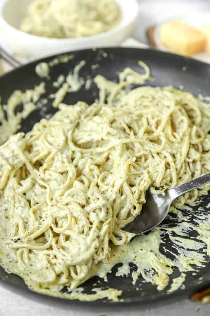 pesto alfredo in a black non-stick pan with a silver spoon scooping.