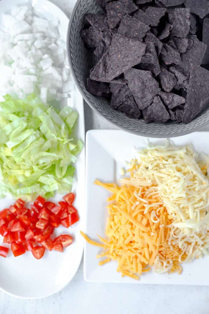 overhead photo of the taco dip ingredients: white onions, chopped lettuce, diced tomatoes, blue corn chips, shredded cheeses.