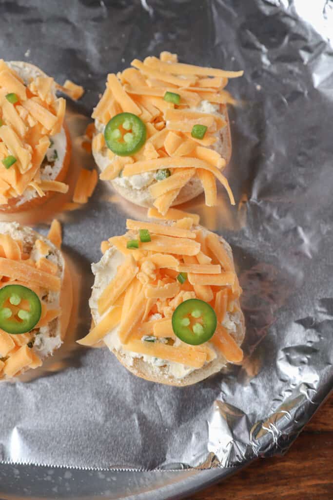 mini bagels on a foil lined baking sheet topped with the cream cheese blend, shredded Cheddar cheese, and jalapeno  slices.