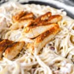 slices of cooked seasoned chicken on top of creamy bacon thin spaghetti in a dark nonstick pan.