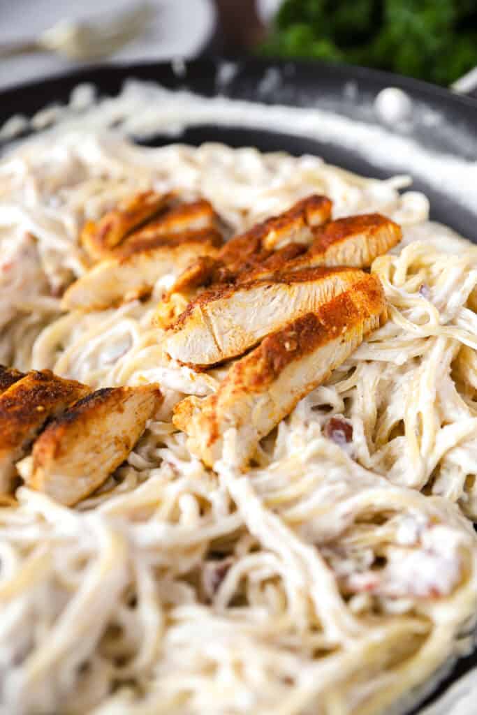 slices of cooked seasoned chicken on top of creamy bacon thin spaghetti in a dark nonstick pan.