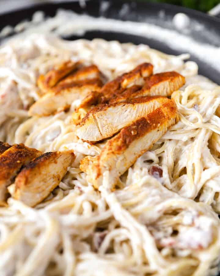 slices of cooked seasoned chicken on top of creamy bacon thin spaghetti in a dark nonstick pan.