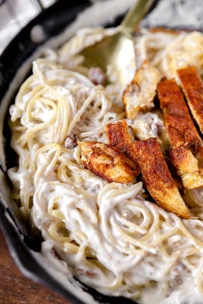 seasoned cooked chicken slices on top of the creamy pasta noodles in the nonstick pan with a gold serving spoon.