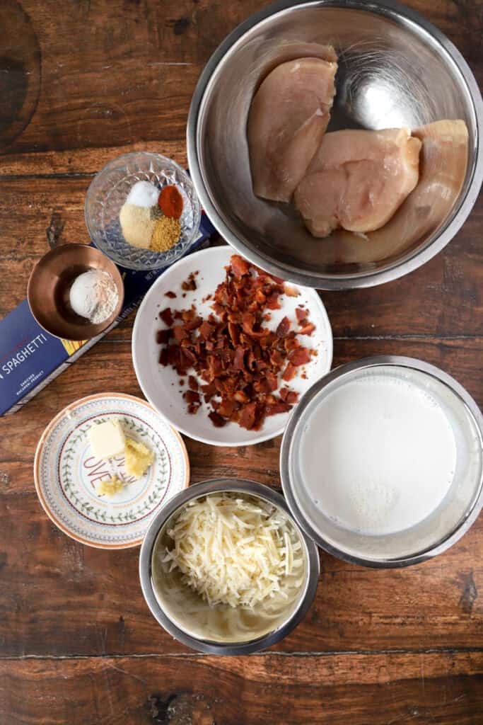 overhead photo of the ingredients on a wood surface. 2 chicken breasts in a stainless steel bowl, seasonings in a glass bowl, flour in a copper dish, box of spaghetti, crumbled bacon on a white plate, butter and grated garlic on a plate, shredded cheeses in a stainless steel bowl, cream in a stainless steel bowl.