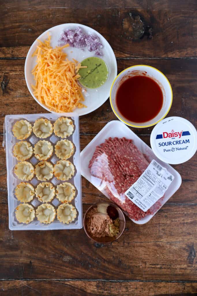 overhead photo of the ingredients on a wood surface: cheese, diced red onions, guac, phyllo cups, ground beef, tomato paste with water, sour cream, seasonings in a small copper dish.