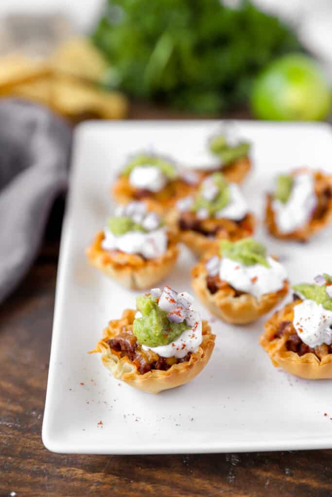 taco bites on a white platter with tortilla chips, parsley, and limes blurred in the background.