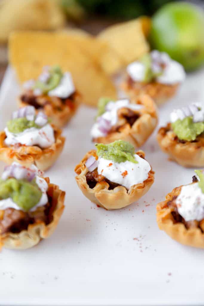 taco bites in phyllo cups on a white platter with tortilla chips and a lime in the background.