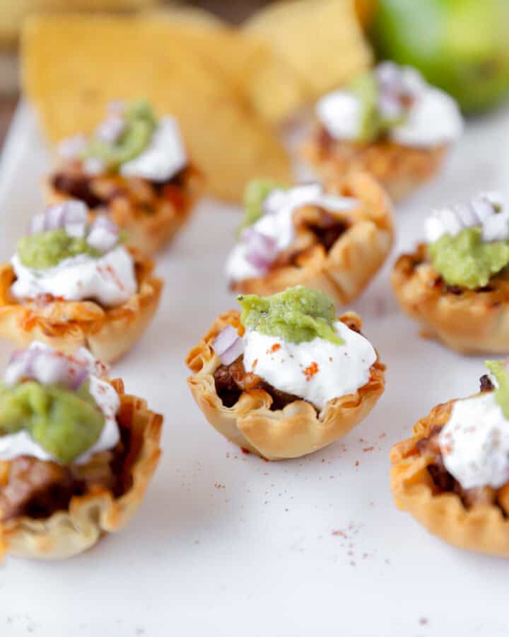 taco bites in phyllo cups on a white platter with tortilla chips and a lime in the background.