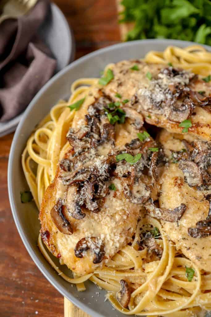 chicken breast marsala covered in marsala mushrooms on a bed of fettucine noodles on a grey round plate. A bunch of parsley in the background.