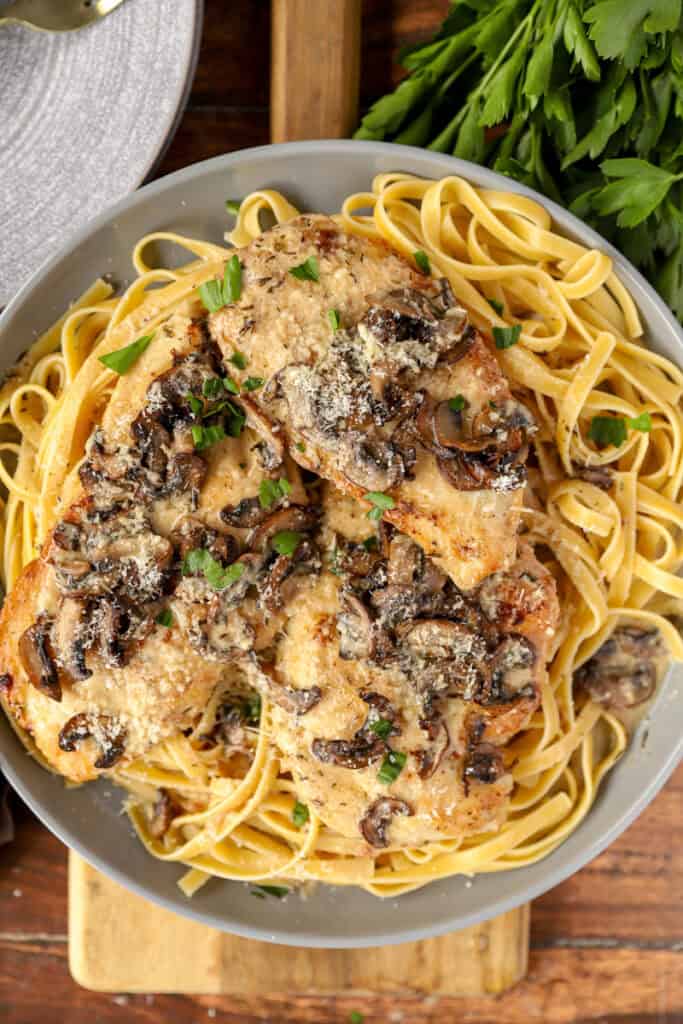 overhead photo of the cooked chicken breasts smothered in marsala mushrooms on a bed of fettucine noodles on a grey plate.