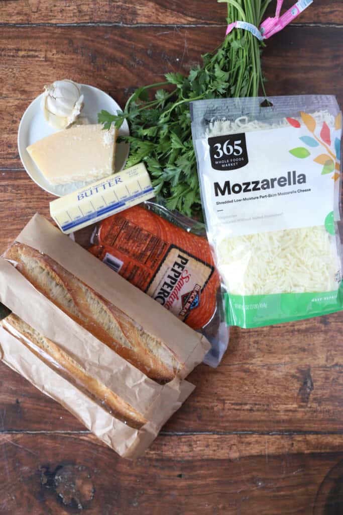 ingredients for the garlic bread on a wood surface: Parmesan, garlic cloves, parsley, shredded Mozzarella, sliced pepperoni, 2 loaves of French bread, one stick of butter