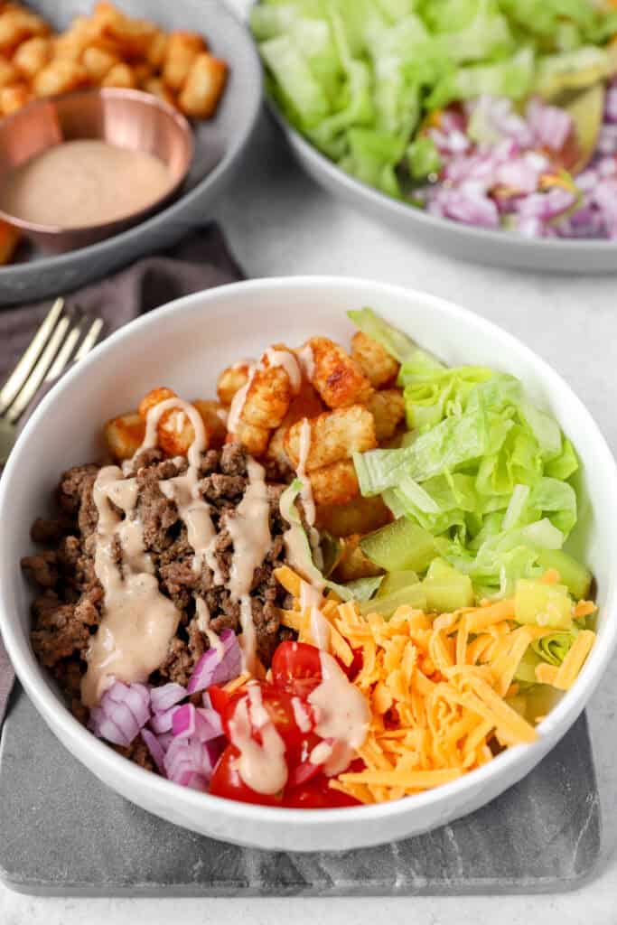the prepared burger bowl ingredients in a round with bowl topped with the special sauce. Ingredients and tater tots blurred in the background.