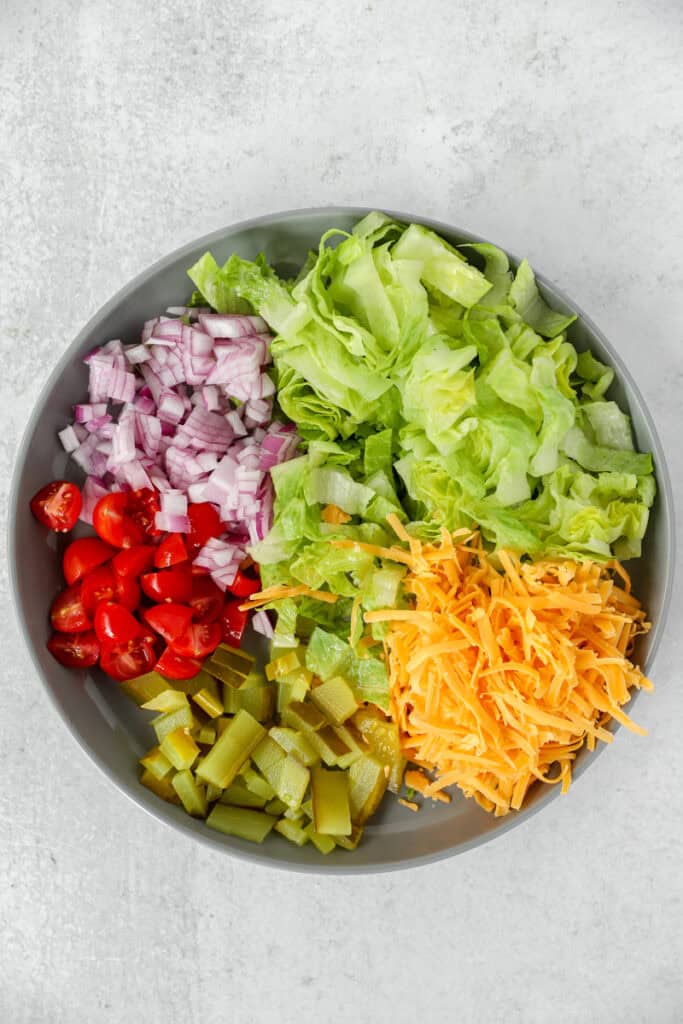 overhead photo of chopped ingredients on a grey round plate: tomatoes, red onions, pickles, lettuce, Cheddar cheese.