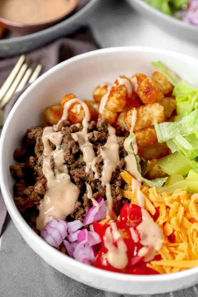 close photo of the burger bowls showing the cooked ground beef topped with the sauce in a round white bowl. Also showing diced red onions, tomatoes, tater tots, lettuce, pickles, and shredded Cheddar cheese.