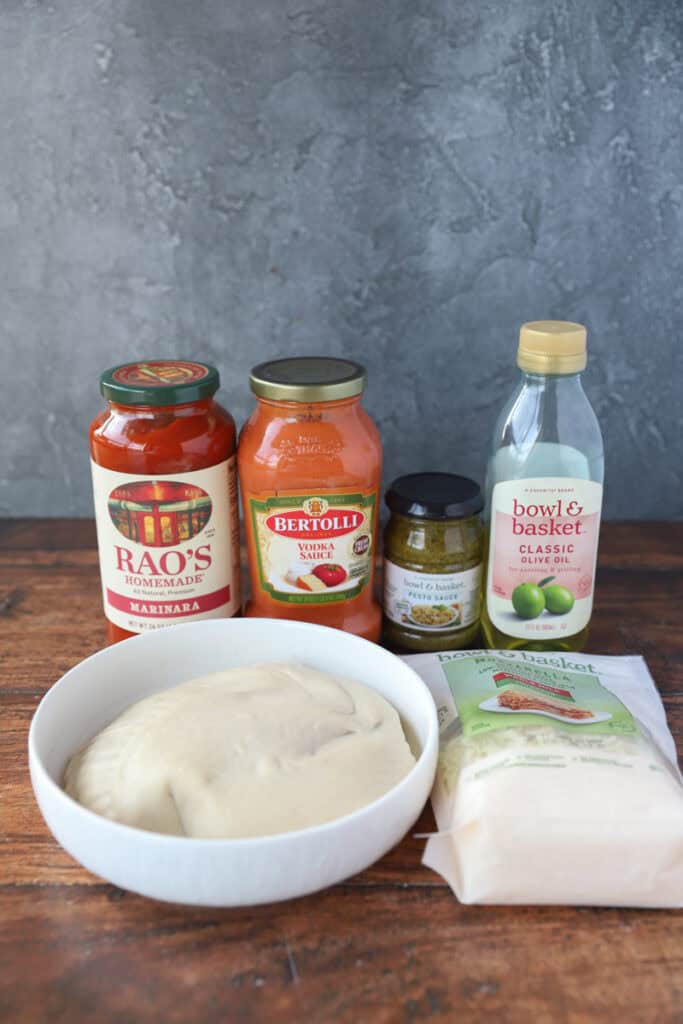 photo of ingredients: jars of marinara, vodka sauce, pesto, olive oil, fresh pizza dough in a white bowl, bag of shredded Mozzarella cheese.