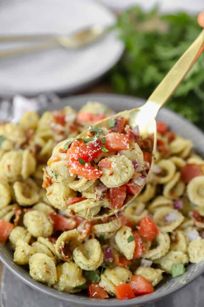 a large gold serving spoon serving some of the bacon pesto pasta with diced tomatoes on top of the pasta.