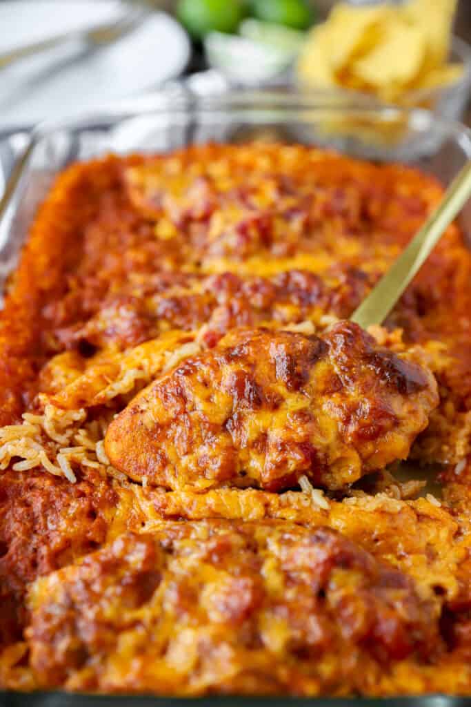a gold serving spoon scooping the salsa chicken from the casserole dish with cooked rice. Tortilla chips, limes, and plates blurred in the background.