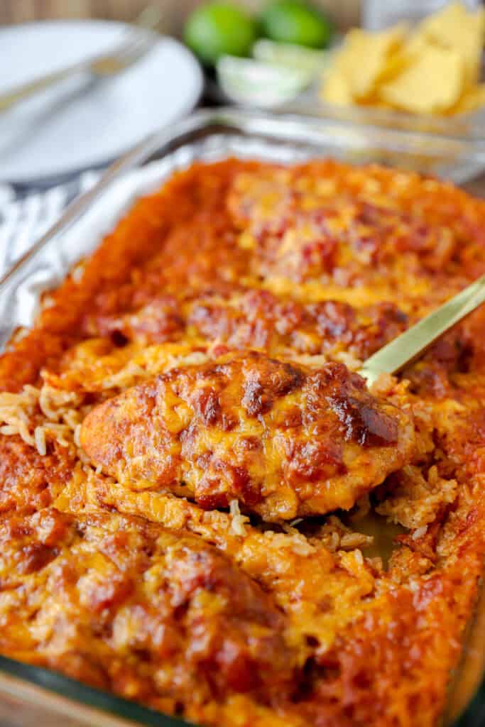 a gold spoon scooping the salsa chicken casserole dish with cooked rice. Tortilla chips, limes, and plates blurred in the background.