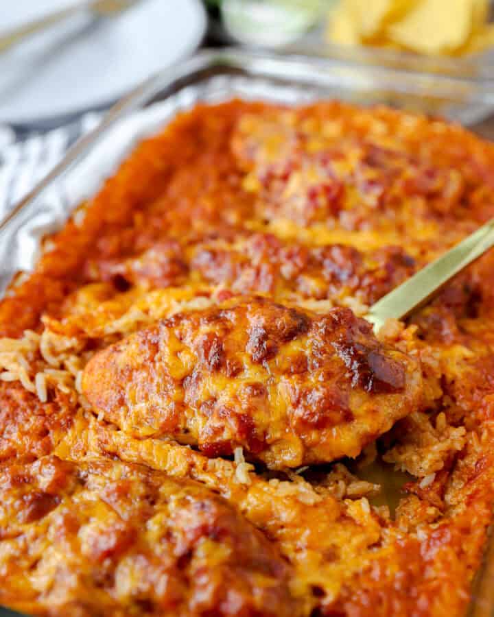 a gold spoon scooping the salsa chicken casserole dish with cooked rice. Tortilla chips, limes, and plates blurred in the background.