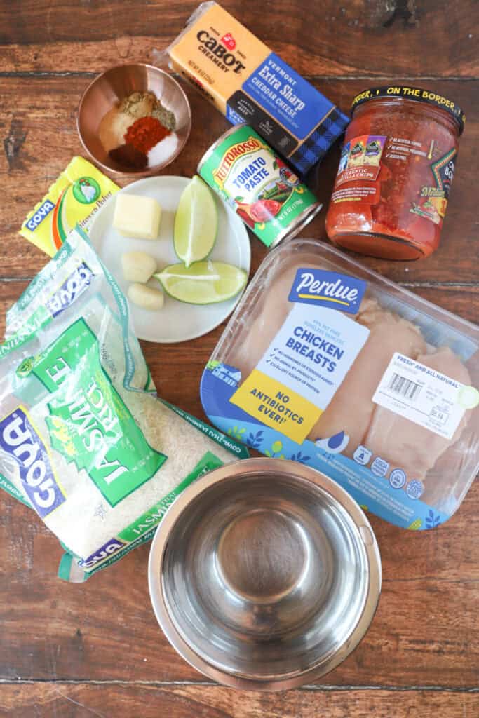 overhead of ingredients flat lay on a word surface: jasmine rice bag, pack of chicken breasts, stainless steel bowl of water, limes, garlic cloves, butter, seasonings, tomato paste, salsa, Cheddar cheese.
