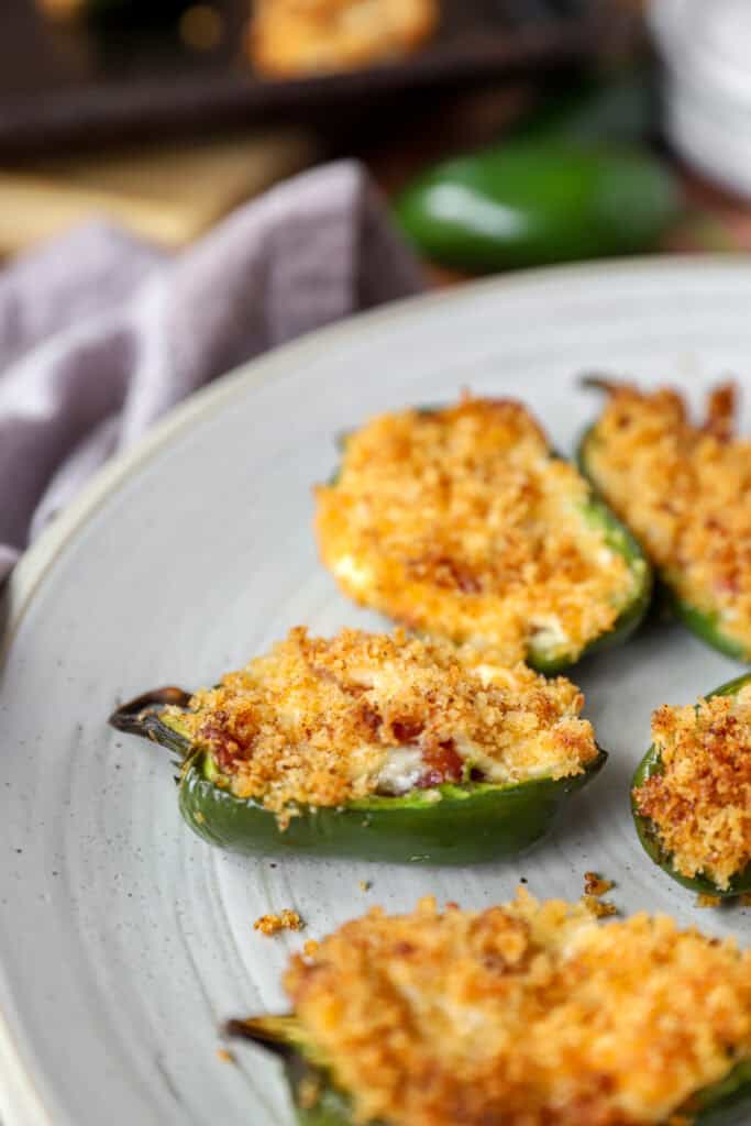 baked jalapeño poppers on a round grey plate. A grey napkin and jalapeño pepper blurred in the background.