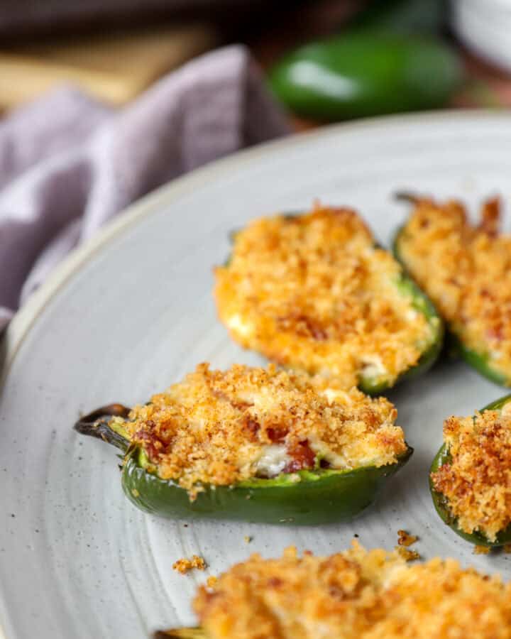 baked jalapeño poppers on a round grey plate. A grey napkin and jalapeño pepper blurred in the background.