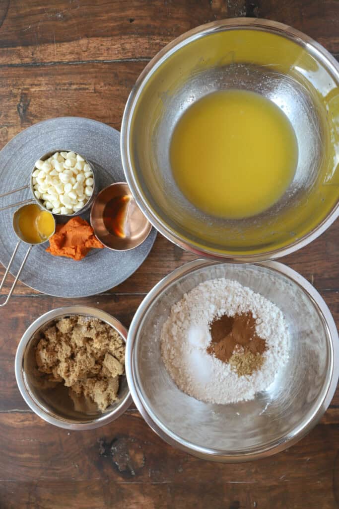overhead flat lay photo of the ingredients on a wood surface. White chocolate chips, egg yolk, vanilla, pumpkin, melted butter, brown sugar, flour, spices, salt, and baking soda.