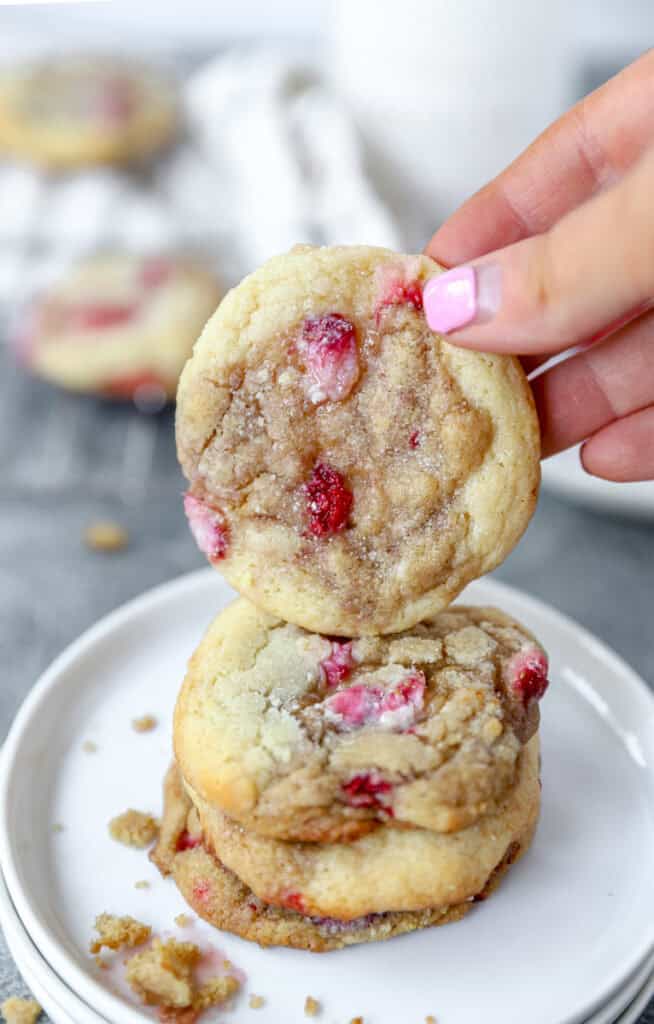 a hand holding one of the strawberry and cream cookies over a stack of cookies on a round white plate.