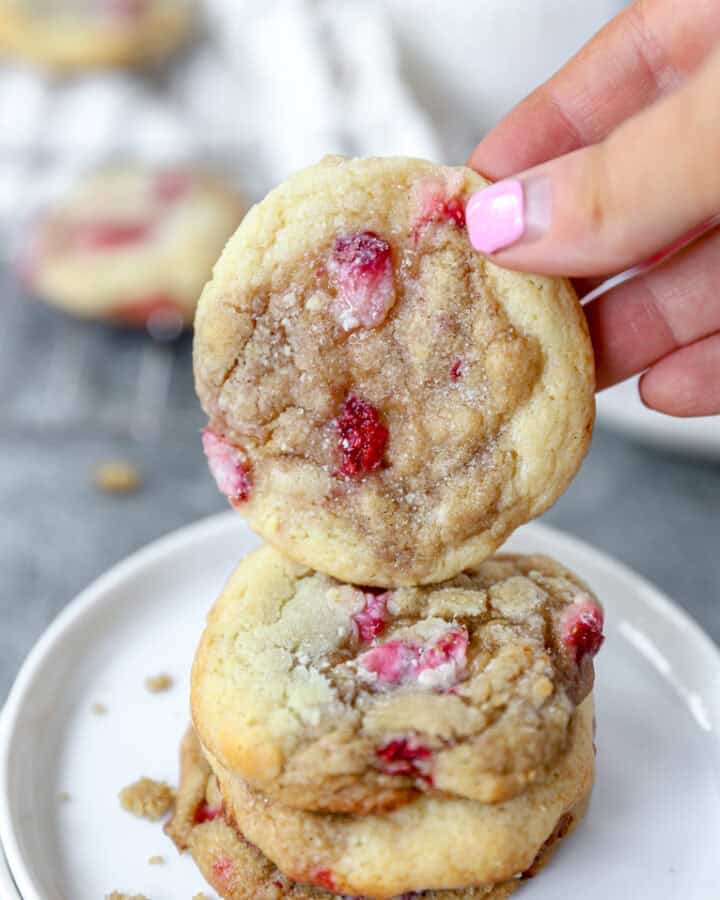 a hand holding one of the strawberry and cream cookies over a stack of cookies on a round white plate.