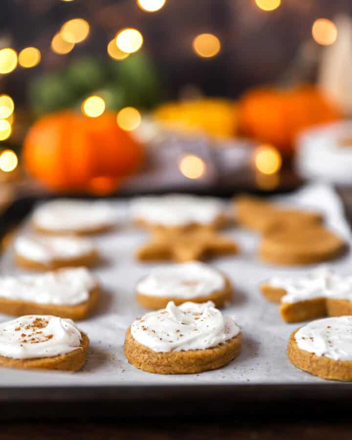 baked and frosted pumpkin sugar cookies on a parchment paper lined baking sheet. Blurred white string lights and pumpkins in the background.