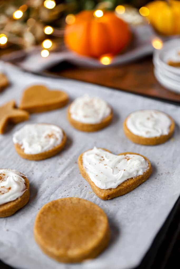 baked and frosted heart shaped and circle pumpkin sugar cookies on a parchment paper lined baking sheet. Blurred white string lights and pumpkins in the background.