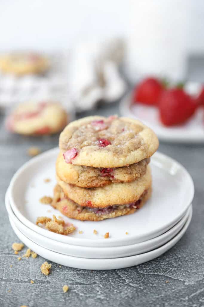 a stack of 4 cookies on a stack of 3 round white plates. Strawberries and more cookies blurred in the background.
