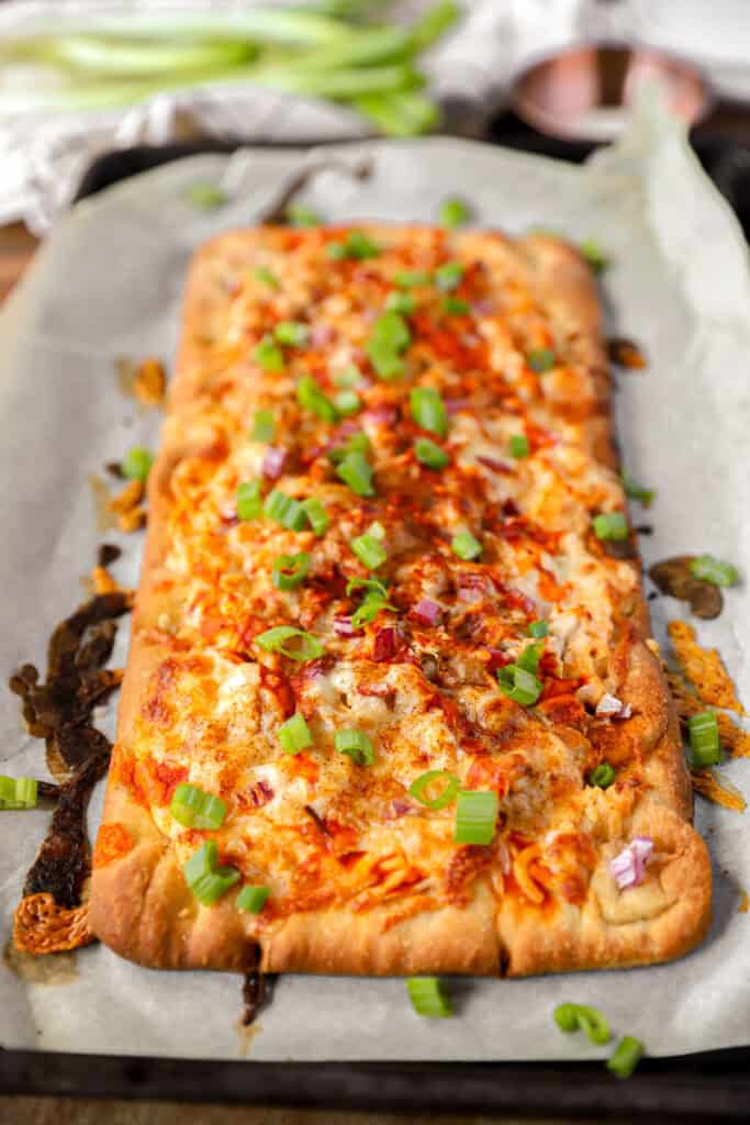 baked Buffalo chicken flatbread topped with diced scallions on parchment paper.
