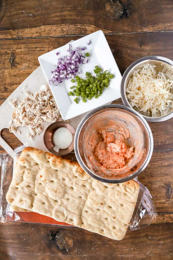 overhead photo of ingredients: diced red onions and scallions, shredded cheeses, chopped rotisserie chicken, flatbread, ranch, cream cheese mixed with hot sauce.