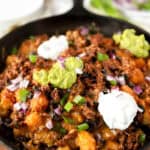 baked totchos topped with guac, sour cream, scallions, and red onions in a cast iron skillet.