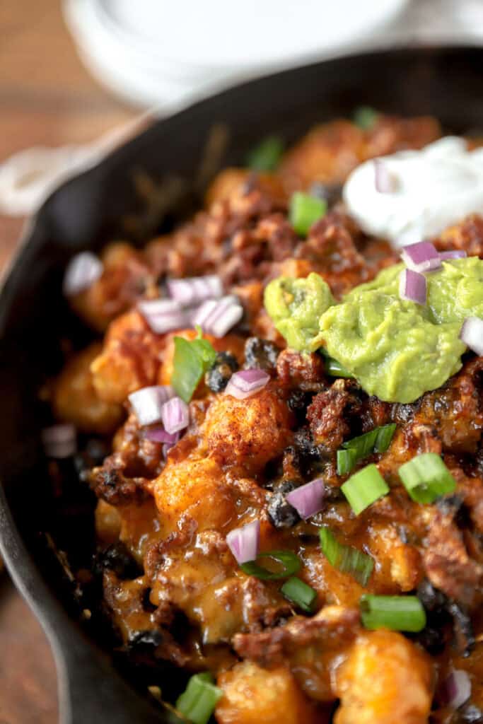 close view of the baked totchos topped with guac, sour cream, scallions, and red onions in a cast iron skillet.