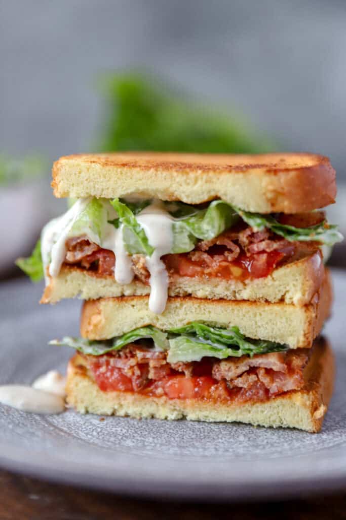 Two pieces Caesar BLT sandwich stacked on top of each other on a grey round plate. Caesar dressing is dripping from the top piece.