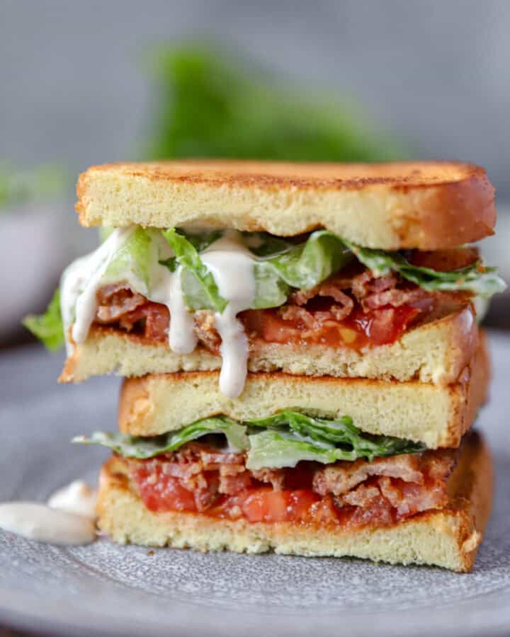 Two pieces Caesar BLT sandwich stacked on top of each other on a grey round plate. Caesar dressing is dripping from the top piece.