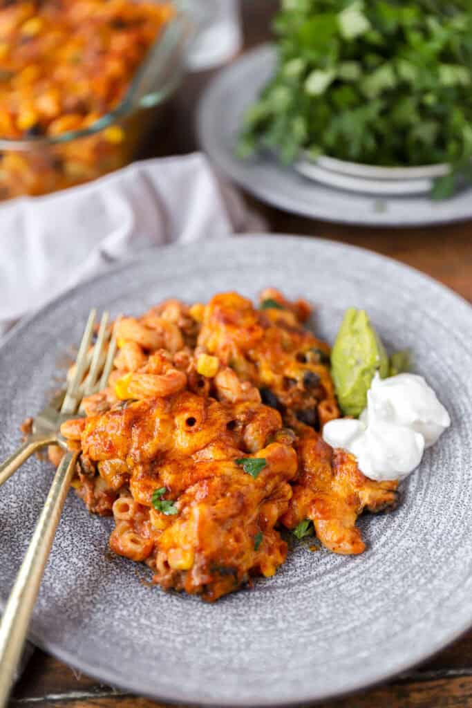 a serving of the taco pasta bake with sour cream and guacamole on a grey round plate with two gold forks.