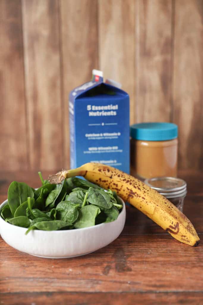ingredients: fresh spinach leaves in a white bowl, banana, almond milk, peanut butter, cinnamon.