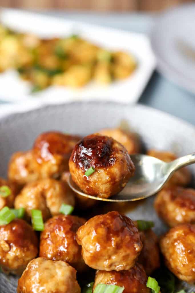 a spoon holding one of the meatballs over a grey bowl of the rest of the baked meatballs.