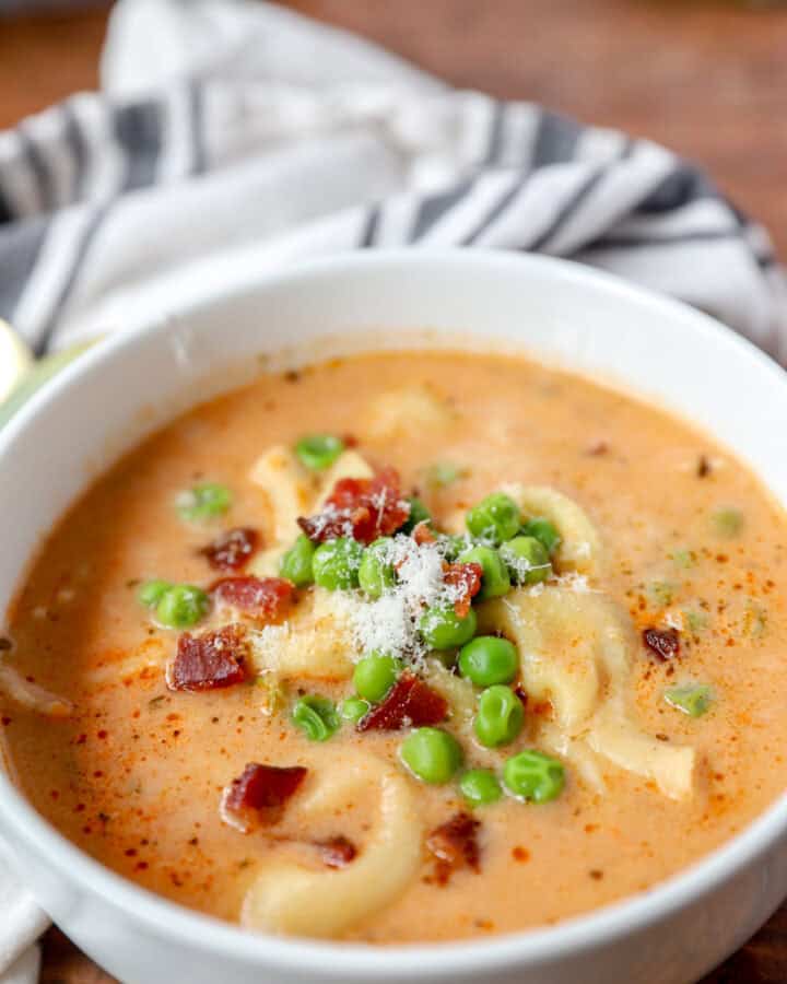 creamy tortellini soup topped with bacon and peas in a round white bowl.